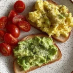 Plate with avocado toast, scrambled egg toast, and fresh cherry tomatoes for a healthy breakfast.