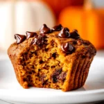 Moist pumpkin chocolate chip muffin with a bite taken out, sitting on a white plate, pumpkins in background