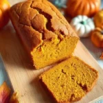 Moist homemade pumpkin bread loaf with a golden crust, sliced to show soft texture, surrounded by pumpkins and autumn leaves.