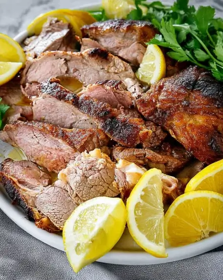 Platter of sliced roasted lamb garnished with lemon wedges and fresh parsley on a gray linen background.