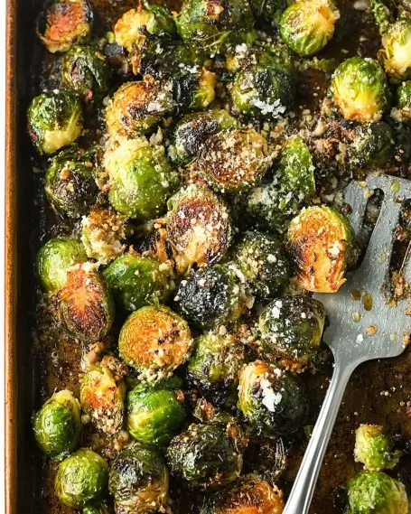 Crispy roasted Brussels sprouts on a baking sheet, topped with parmesan cheese and garlic, with a metal spatula.