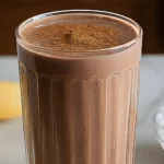 A glass of chocolate smoothie topped with a sprinkle of cocoa powder, with a side of peanut butter and banana.