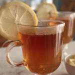 A close-up of a cup of tea with a lemon slice on the rim, with another cup of tea in the background and a bowl of lemon slices in the foreground.