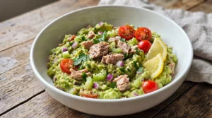 Creamy Avocado Tuna Salad with tuna, mashed avocado, cherry tomatoes, cucumber, red onion, fresh parsley, lemon wedges, and black pepper in a healthy bowl.